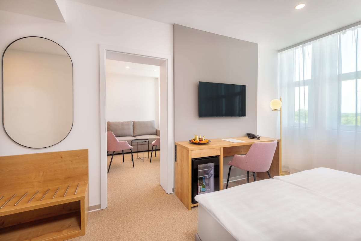 Quarto de hotel moderno com cama, escrivaninha com computador, TV, mini-frigorífico e área de estar com sofá e cadeiras.