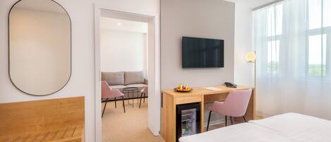 Et moderne hotelværelse med seng, skrivebord med computer, fjernsyn, lille køleskab og siddeområde med sofa og stole.