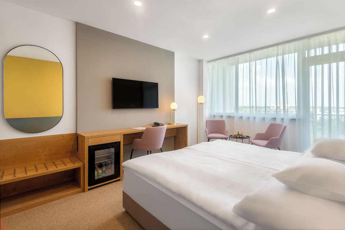 Quarto de hotel com cama, escrivaninha com cadeira, televisão, minibar e uma grande janela com cortinas leves.