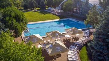 Vue aérienne d’un espace piscine avec des chaises longues et des parasols, entouré d’arbres et d’un bâtiment doté d’un toit incurvé.