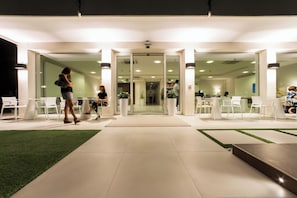 Una entrada moderna a un edificio con puertas de vidrio, sillas blancas y una zona de césped artificial verde.