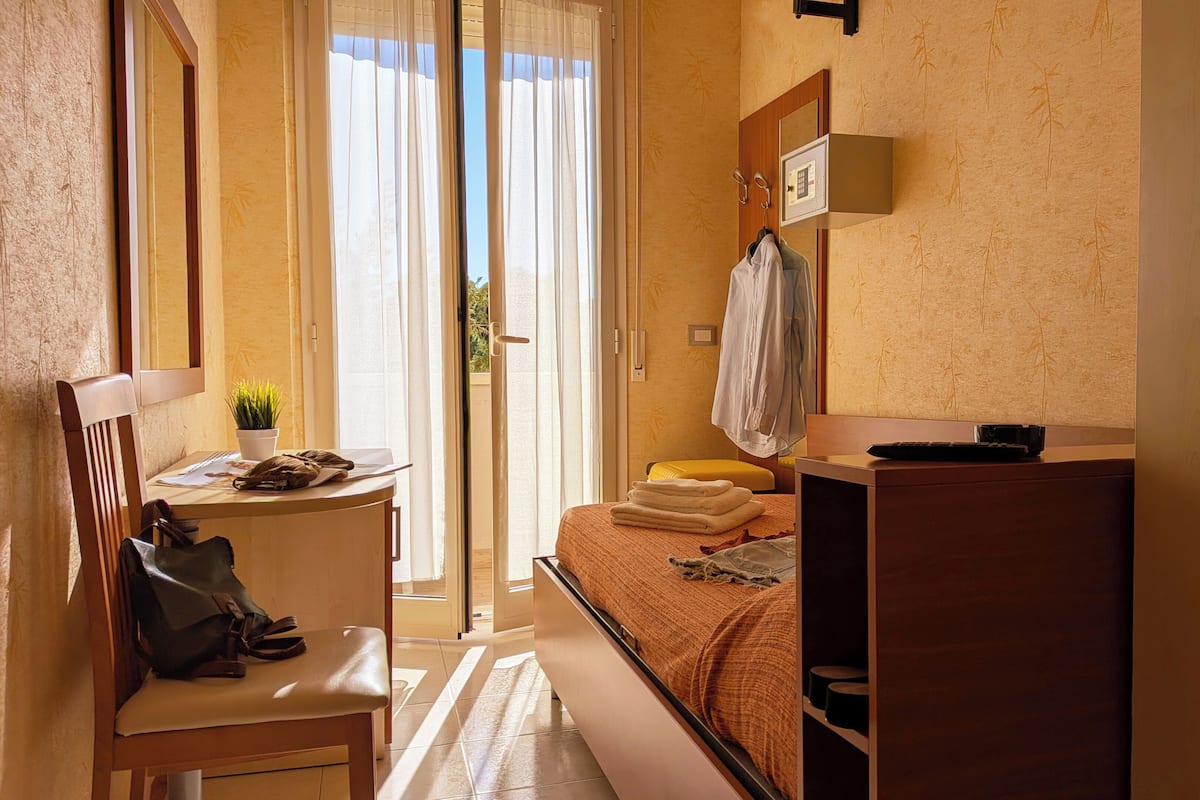 A bedroom with a bed, a desk with a chair, a small table with a plant, and a window with curtains.