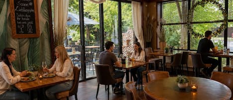 An indoor dining area with wooden tables and chairs, large windows with curtains, and a chalkboard sign.