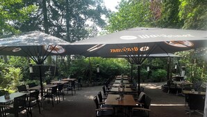 An outdoor dining area with tables and chairs under umbrellas, surrounded by trees. - Hotel-Restaurant Amadeus (Isernhagen)