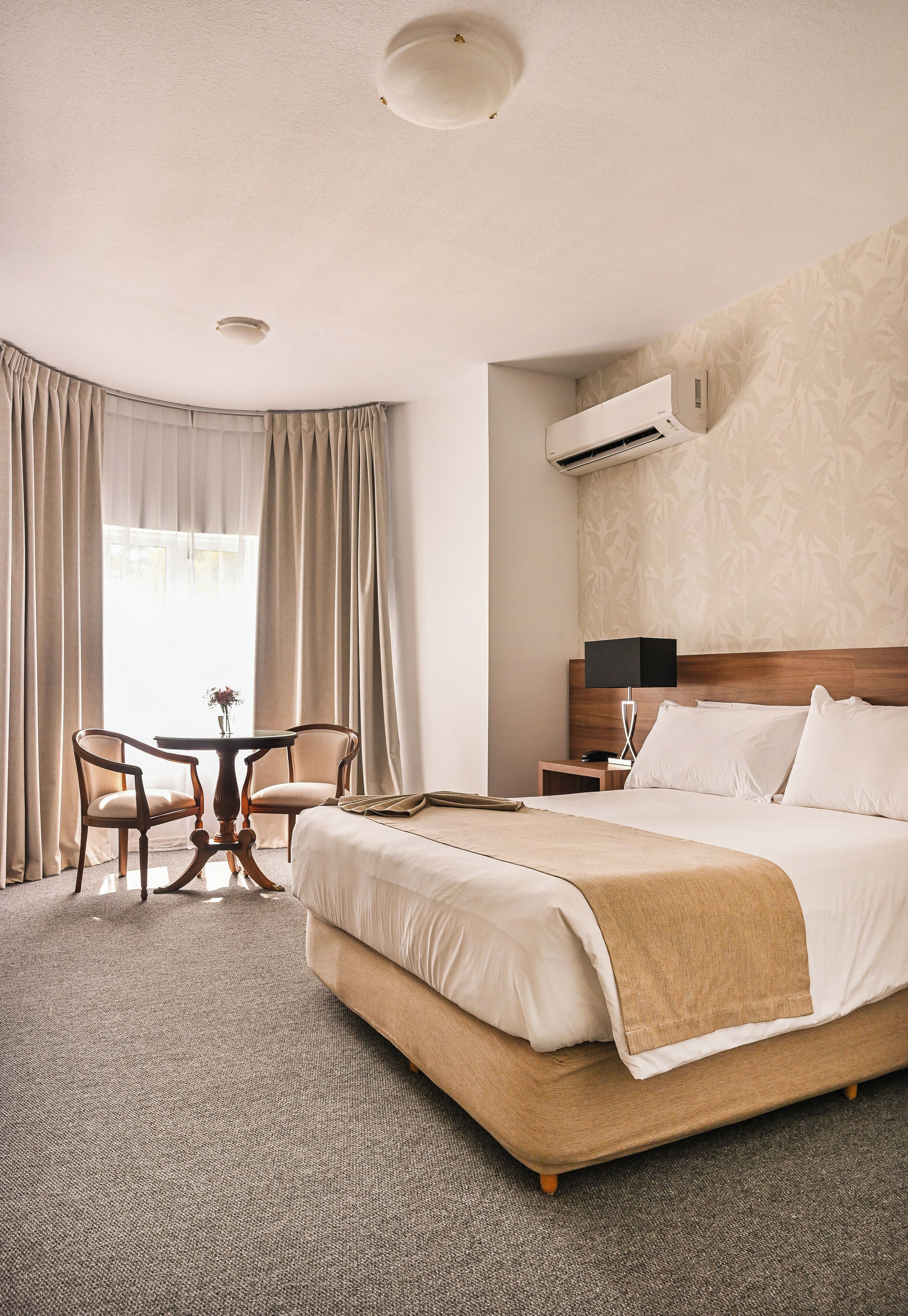 Habitación de hotel con una cama grande, una mesita con dos sillas, una mesita de noche y un equipo de aire acondicionado en la pared.
