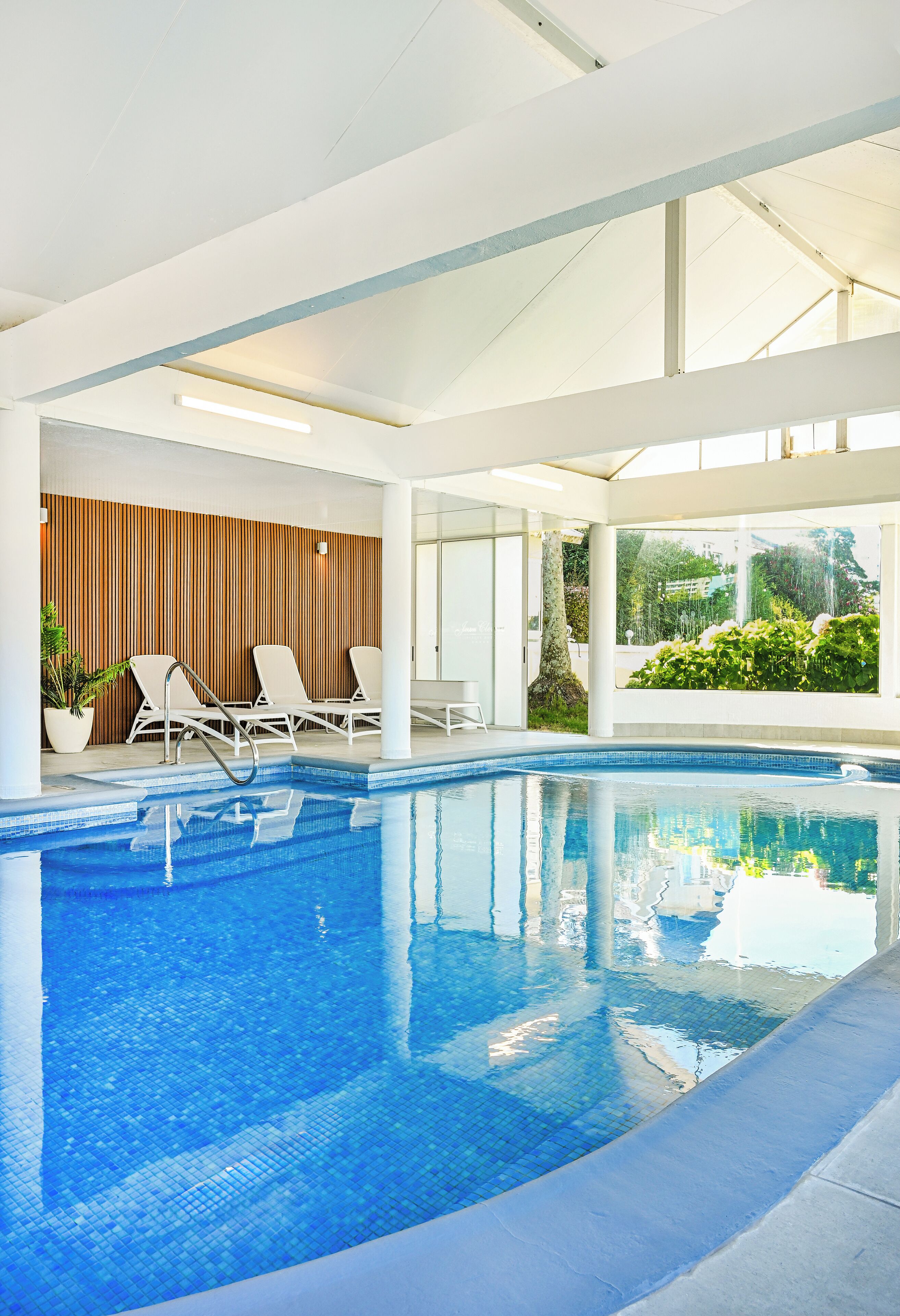 Une piscine intérieure moderne avec une eau d’un bleu cristallin, des chaises longues blanches et un mur en bois avec des lattes verticales.