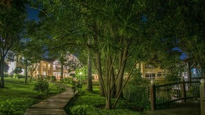A nighttime outdoor pathway with wooden decking, surrounded by lush greenery and trees, leading to a building with multiple windows and balconies. - Parque Hotel Jean Clevers (Punta del Este)