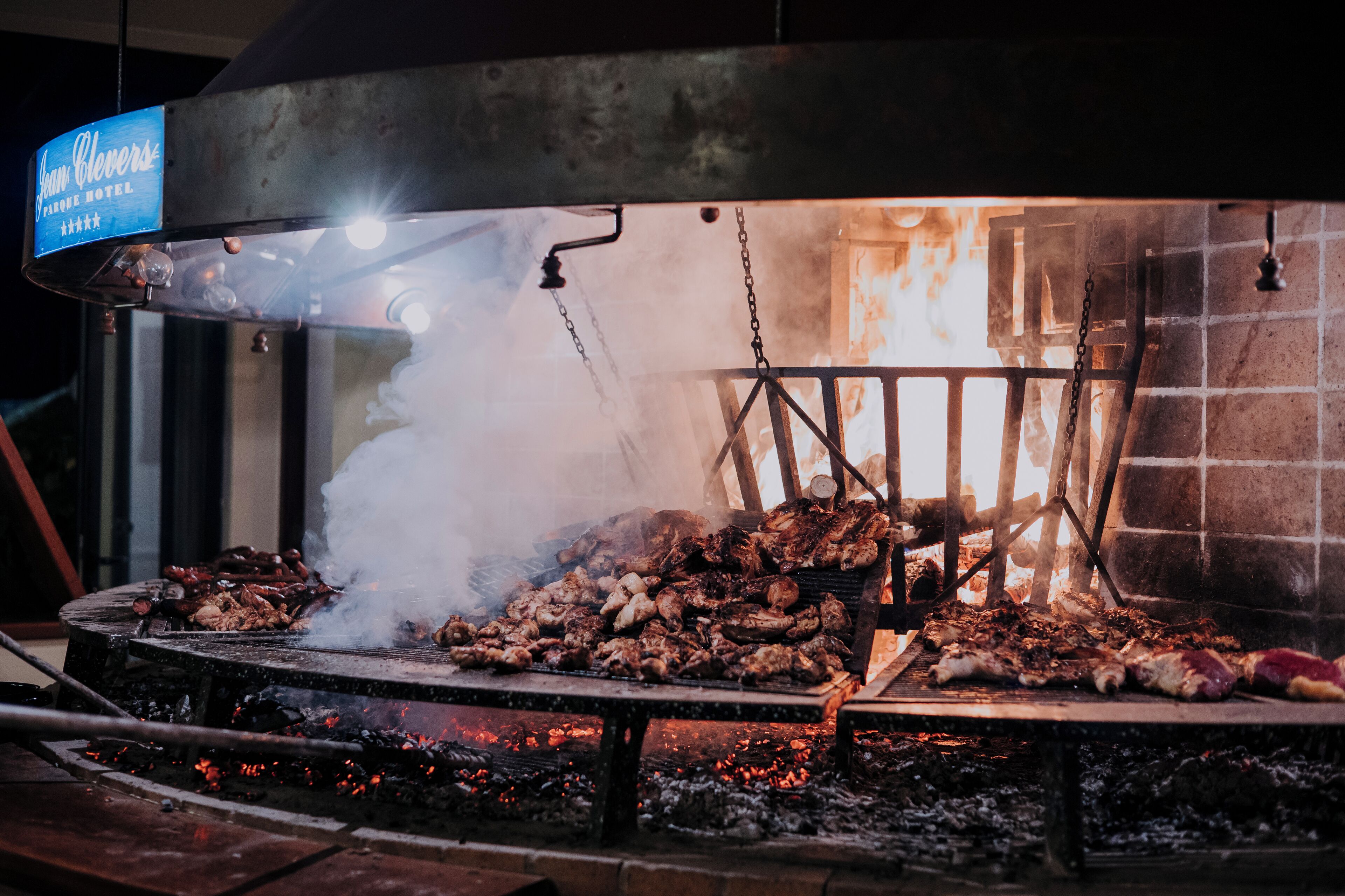 Una parrilla con varios tipos de carne asándose, un cartel encima que indica 'Ian Clevens' y 'Hotel', y un muro de ladrillo al fondo.