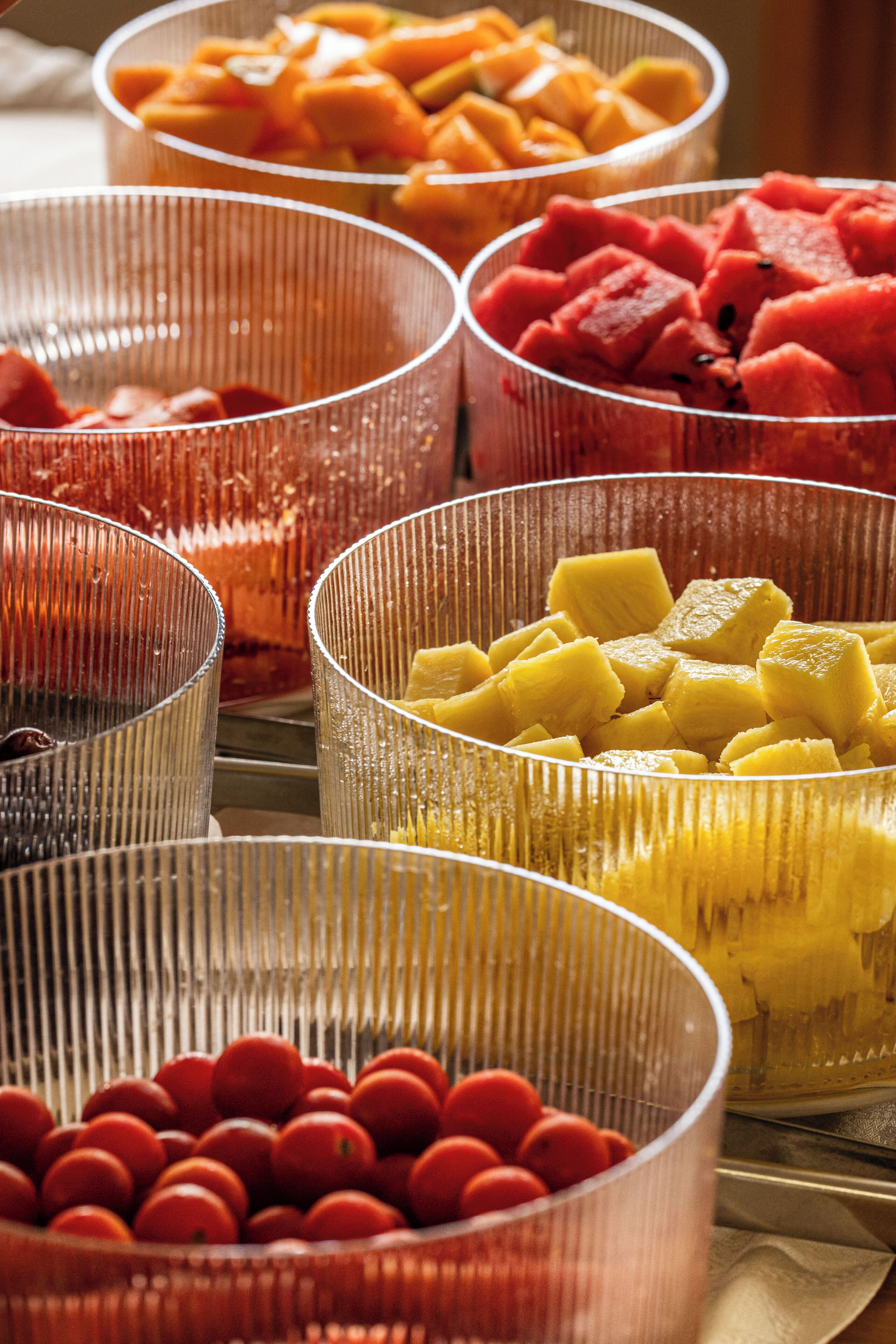 Une variété de fruits présentés dans des bols transparents, notamment des tomates rouges, de l’ananas jaune et de la pastèque rouge.
