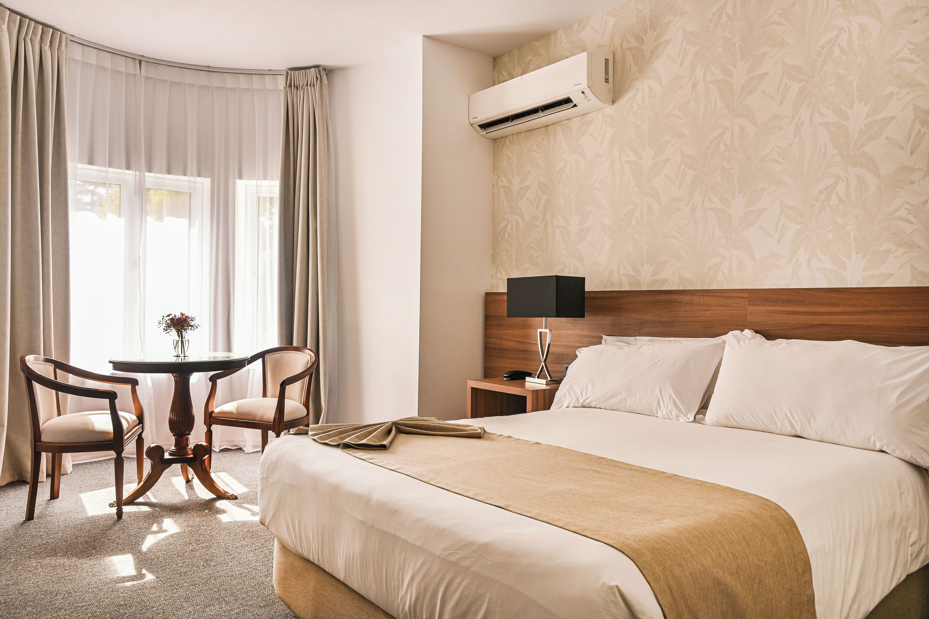 A hotel room with a large bed, a bedside table with a lamp, a small table with chairs, and an air conditioning unit on the wall.