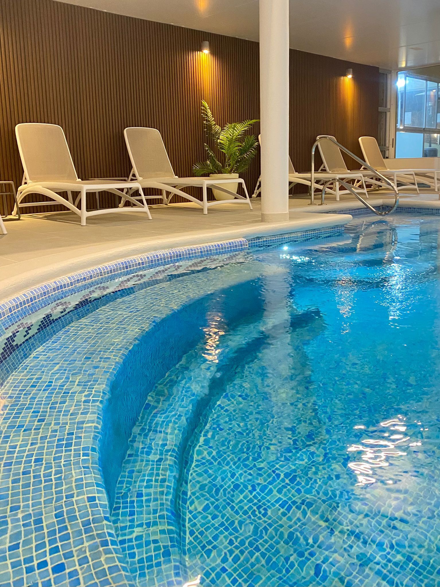 Piscina interior com água azul cristalina, rodeada por espreguiçadeiras brancas e uma parede de madeira com ripas verticais.