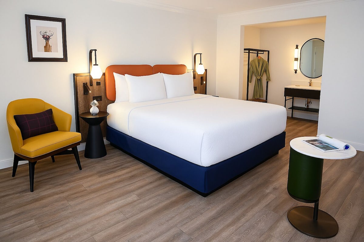 Une chambre d’hôtel comprenant un lit, une chaise, une table d’appoint, une table ronde, un porte-peignoir et un miroir.