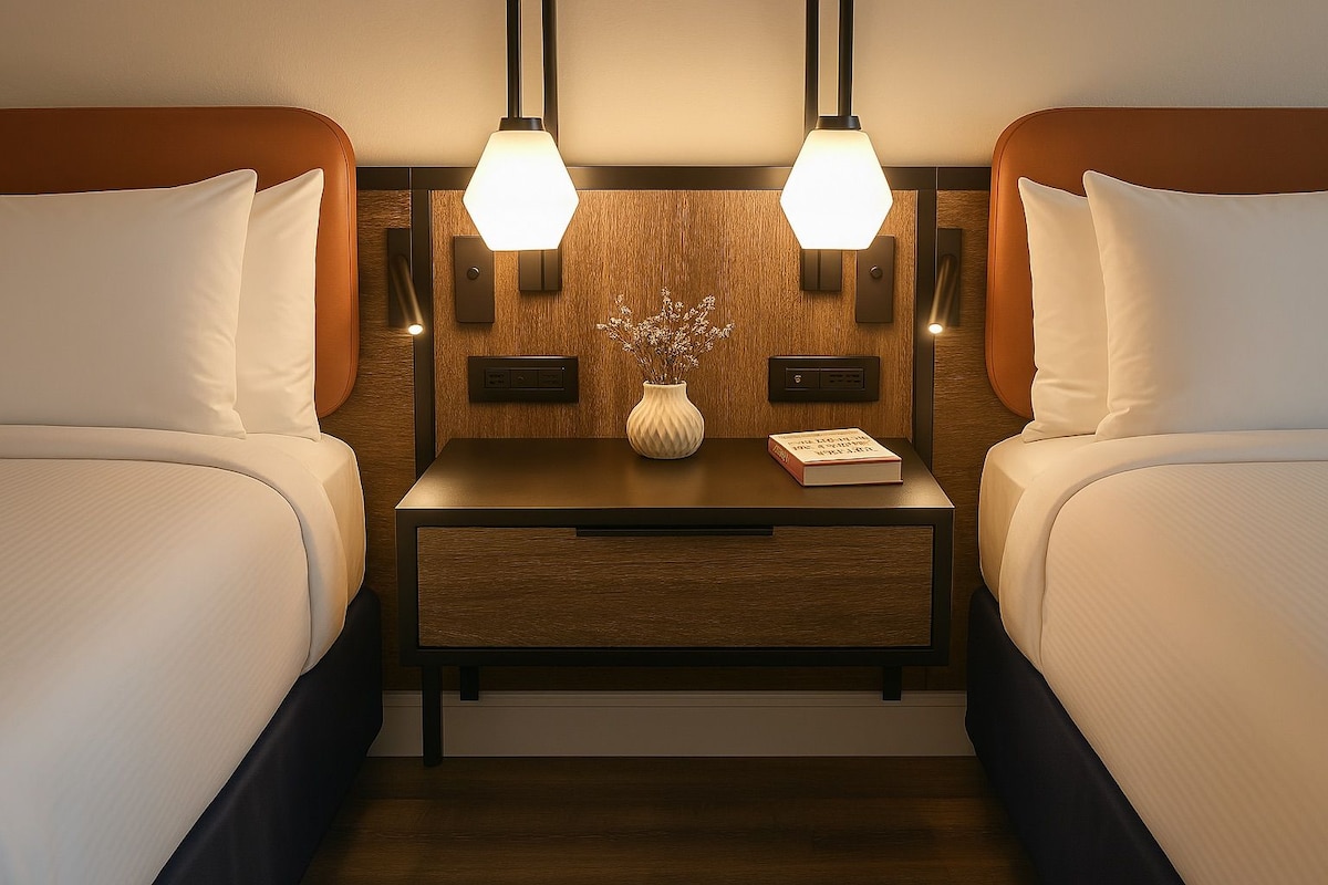 A hotel room with two beds, a nightstand with a vase and a book, and two wall-mounted lamps.