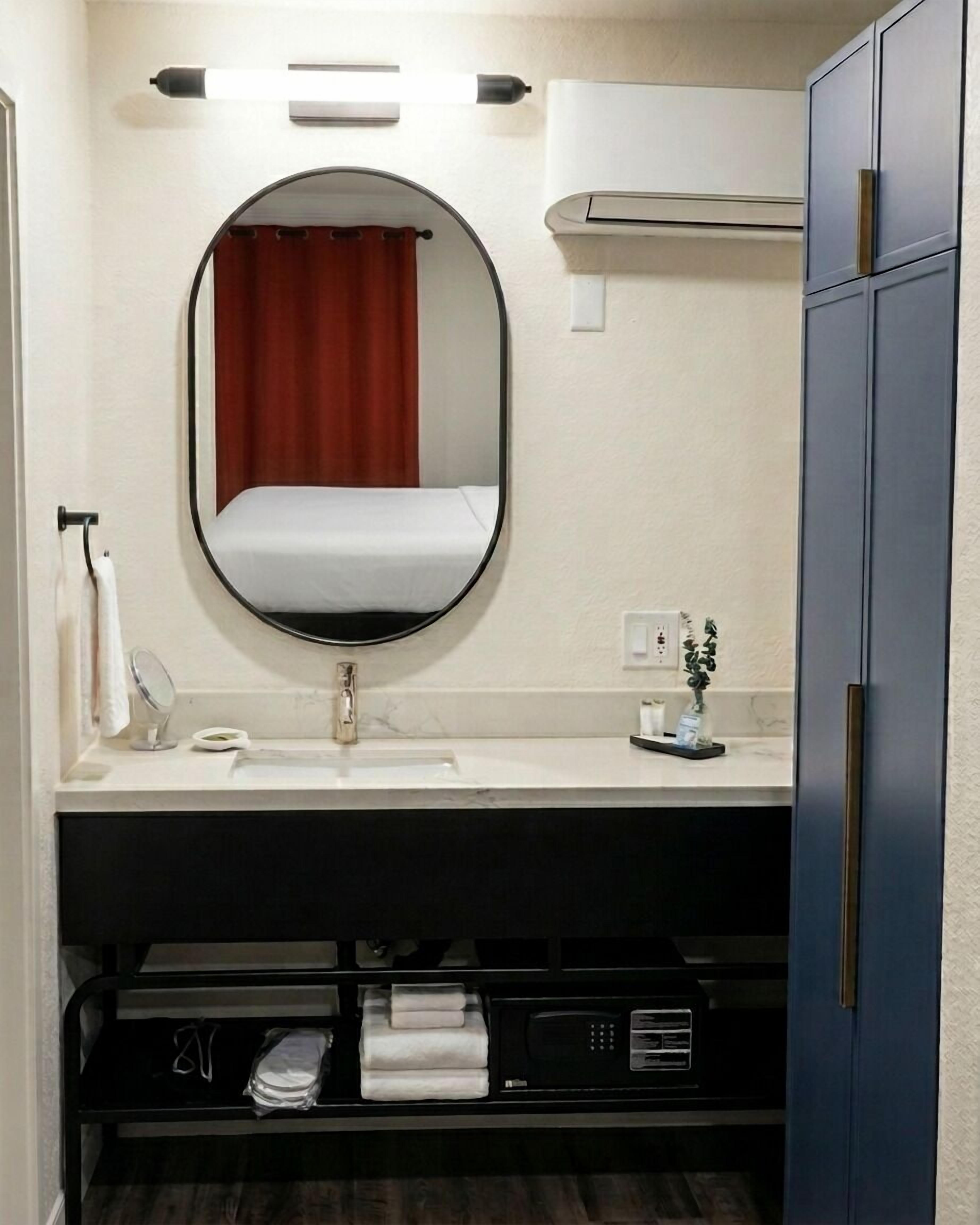A bathroom with a round mirror, a white sink, a red curtain, a towel rack, a small plant, and a cabinet with a microwave.