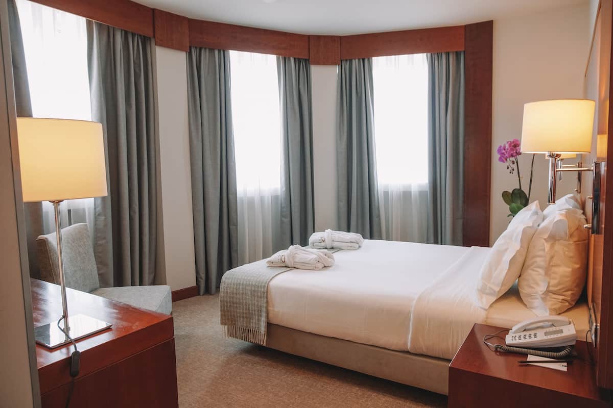 Una habitación de hotel con una cama grande, dos mesitas de noche, un escritorio con lámpara y una ventana con cortinas.