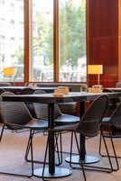 Un restaurante con sillas y mesas de mimbre, amplias ventanas con vistas a edificios y árboles, y una lámpara sobre la mesa.