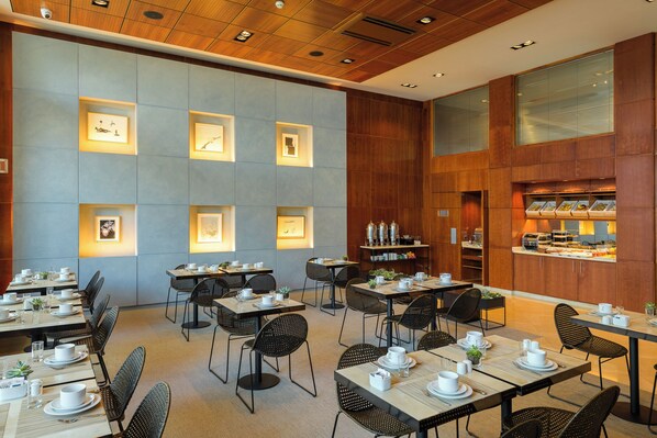 A hotel dining area with multiple tables and chairs, framed artwork on the walls, and a kitchen counter with food displays. - Dom Carlos Liberty Hotel (Lisbon)