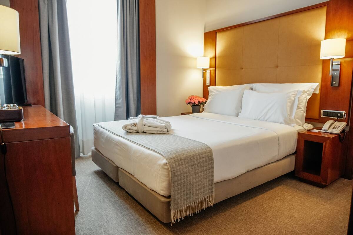 Habitación de hotel con una cama grande, cabecera de madera, mesita de noche con lámpara, teléfono y una pequeña mesa con un florero.