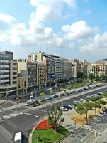 Paisagem urbana com edifícios modernos, uma avenida larga com carros e uma faixa de pedestres.