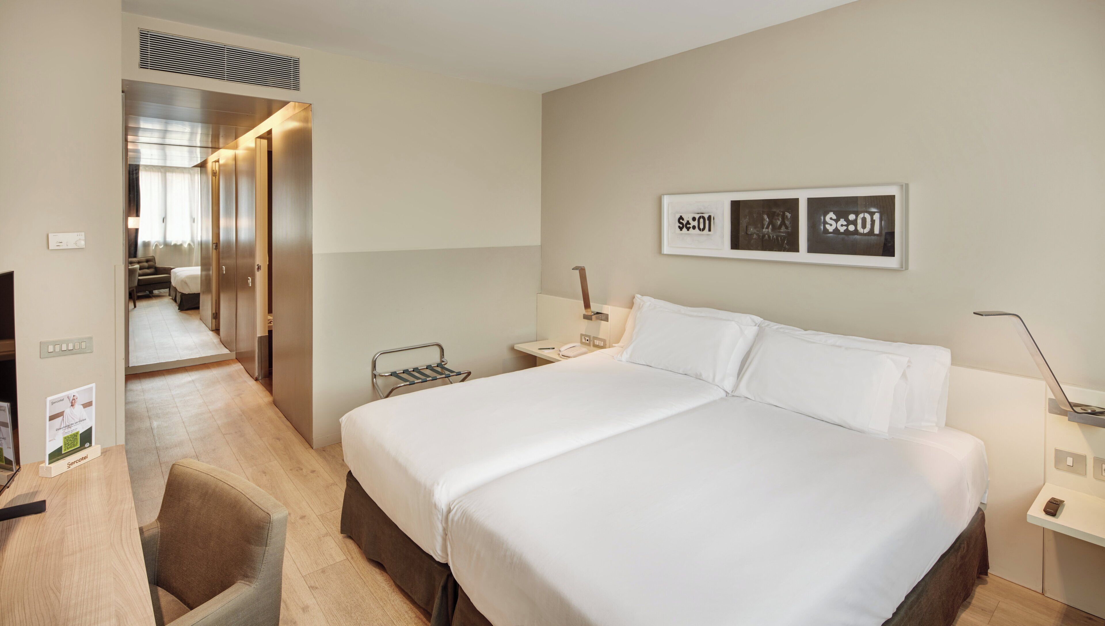 Camera d'albergo con un letto grande, comodini, una scrivania e una sedia. Alle pareti ci sono quadri incorniciati e una bocchetta d'aria sopra la porta.