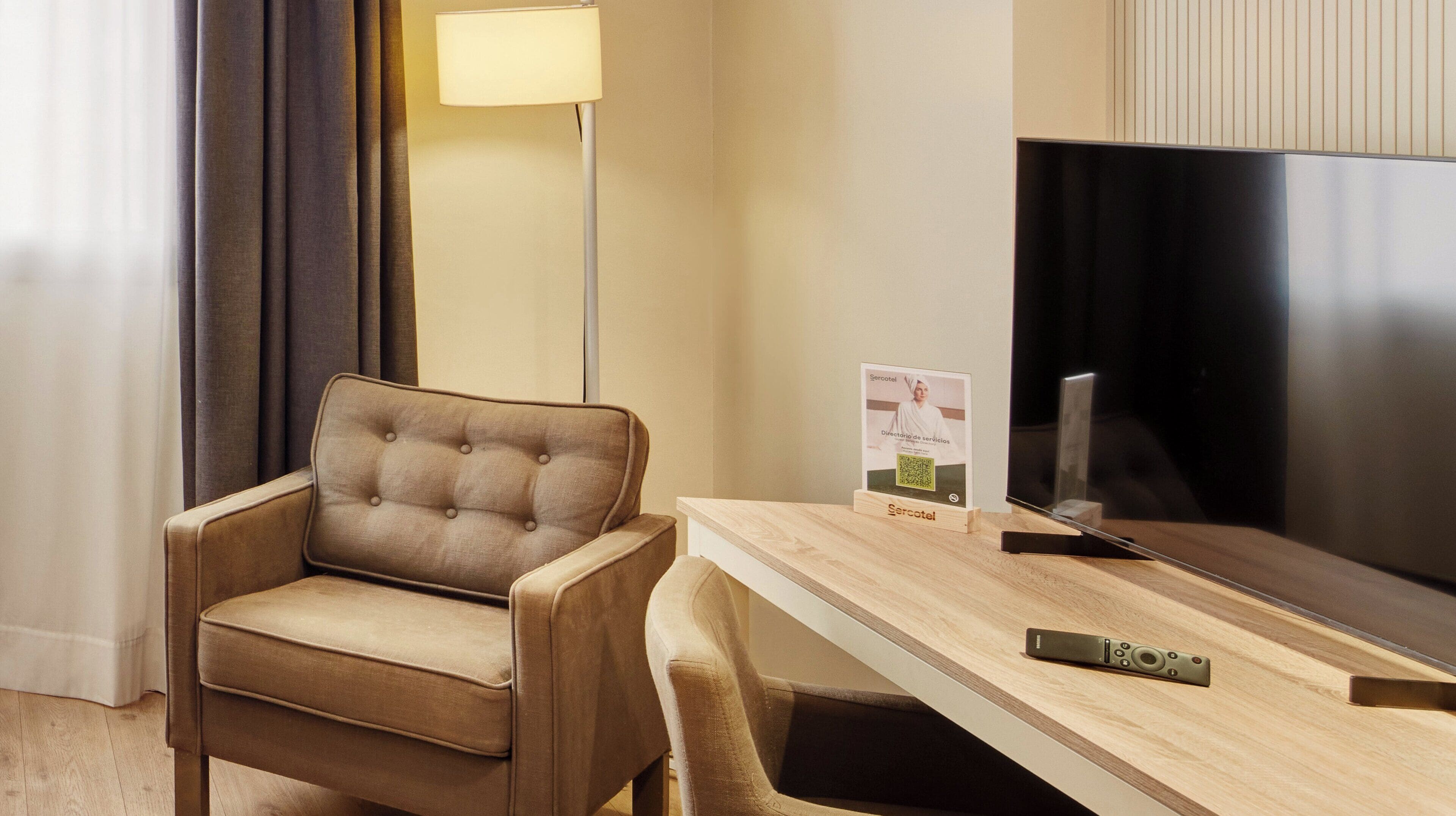 Et hotelværelse med en betrukken lænestol, et træskrivebord, et fladskærms-tv og en lampe.