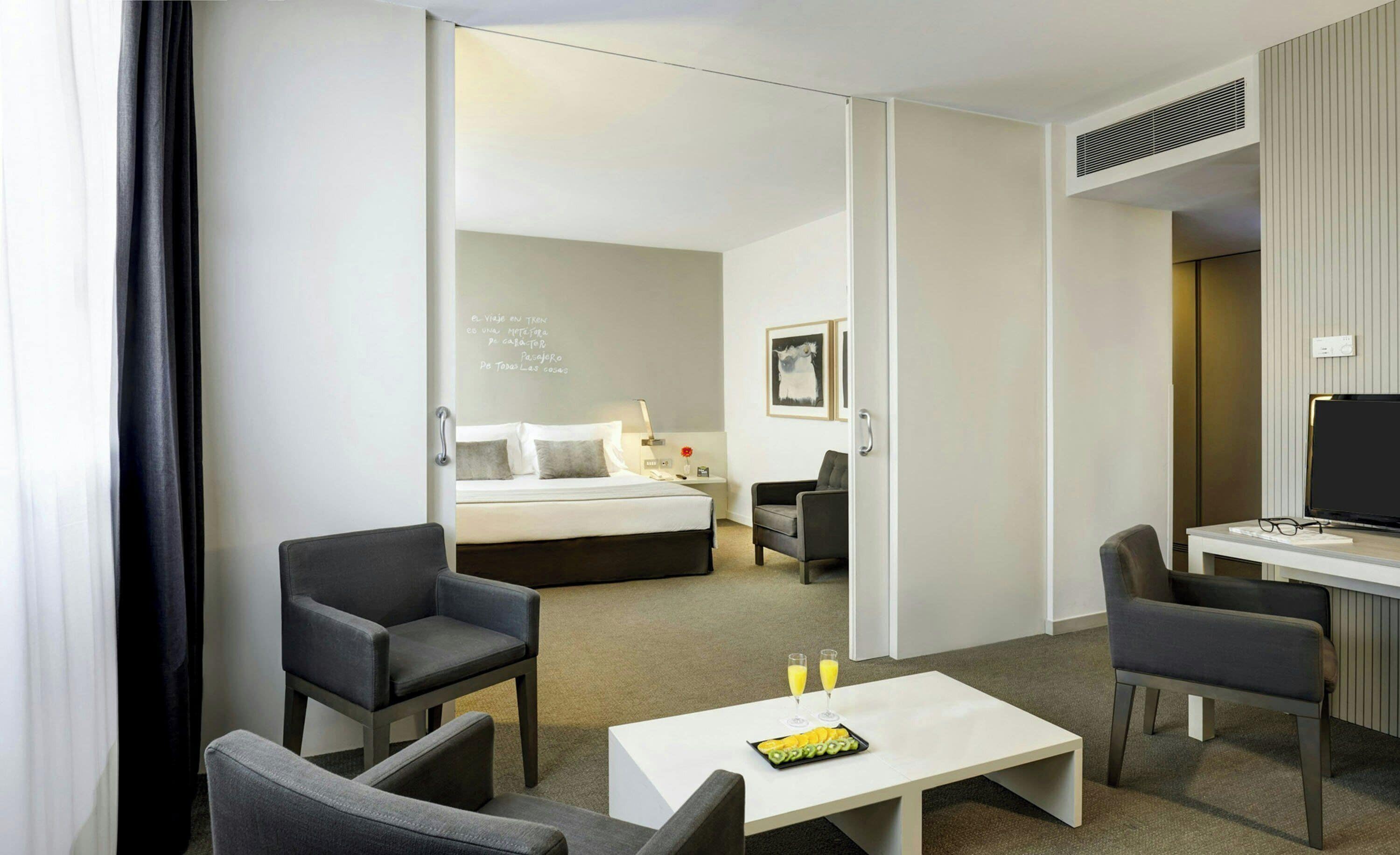 A hotel room with a bed, two armchairs, a small table with a fruit platter, a desk with a computer, and a television.
