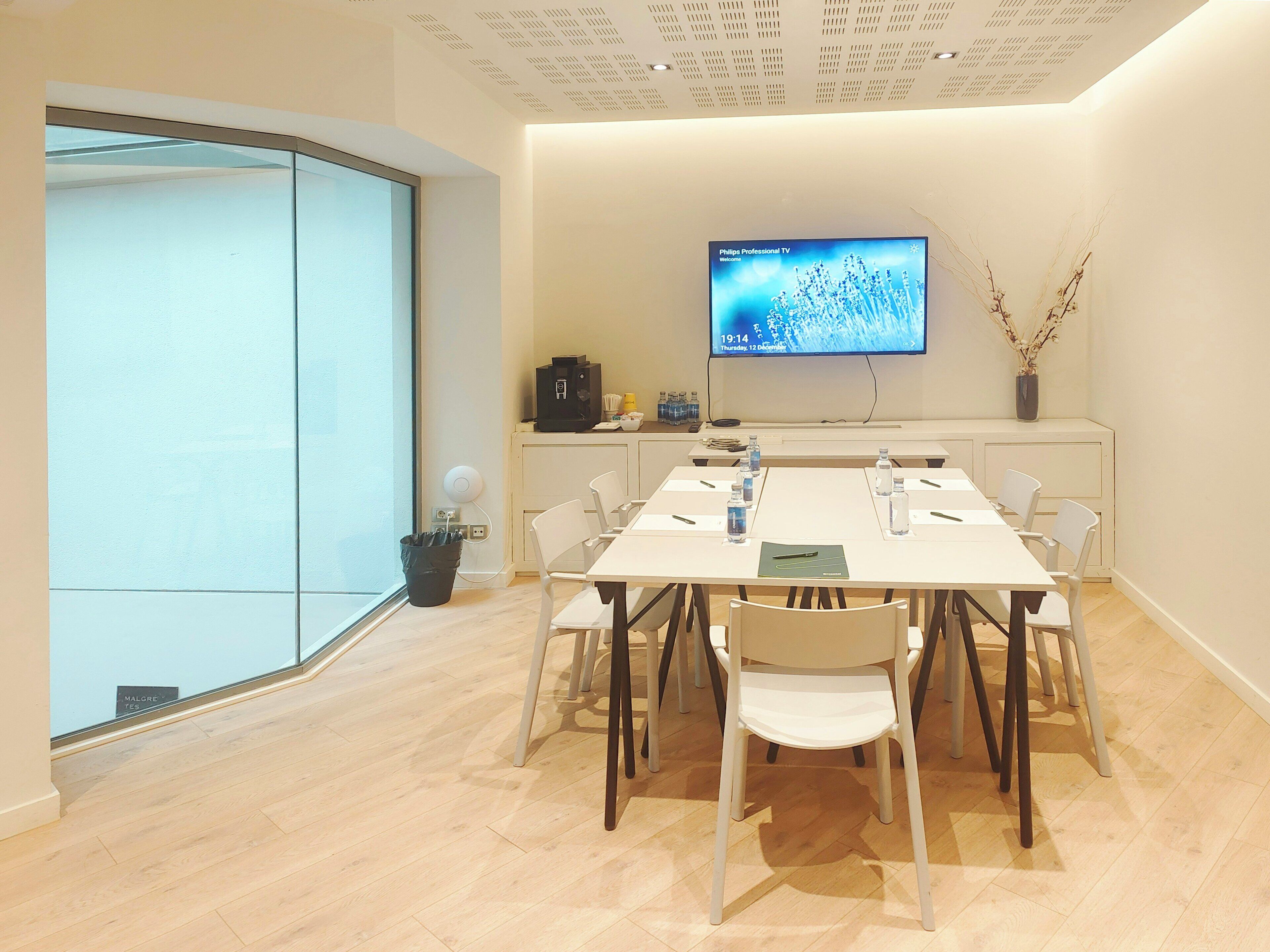 Un salón de conferencias con una mesa larga, sillas blancas, un televisor de pantalla plana, una cafetera y un jarrón con ramas.