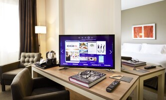 A hotel room with a flat-screen TV displaying a TV channel interface, a bed, a bedside table with a book, glasses, and a remote control.