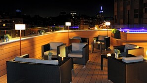 Una terraza en la azotea con mobiliario de mimbre, iluminación ambiental y vistas a la ciudad al fondo.