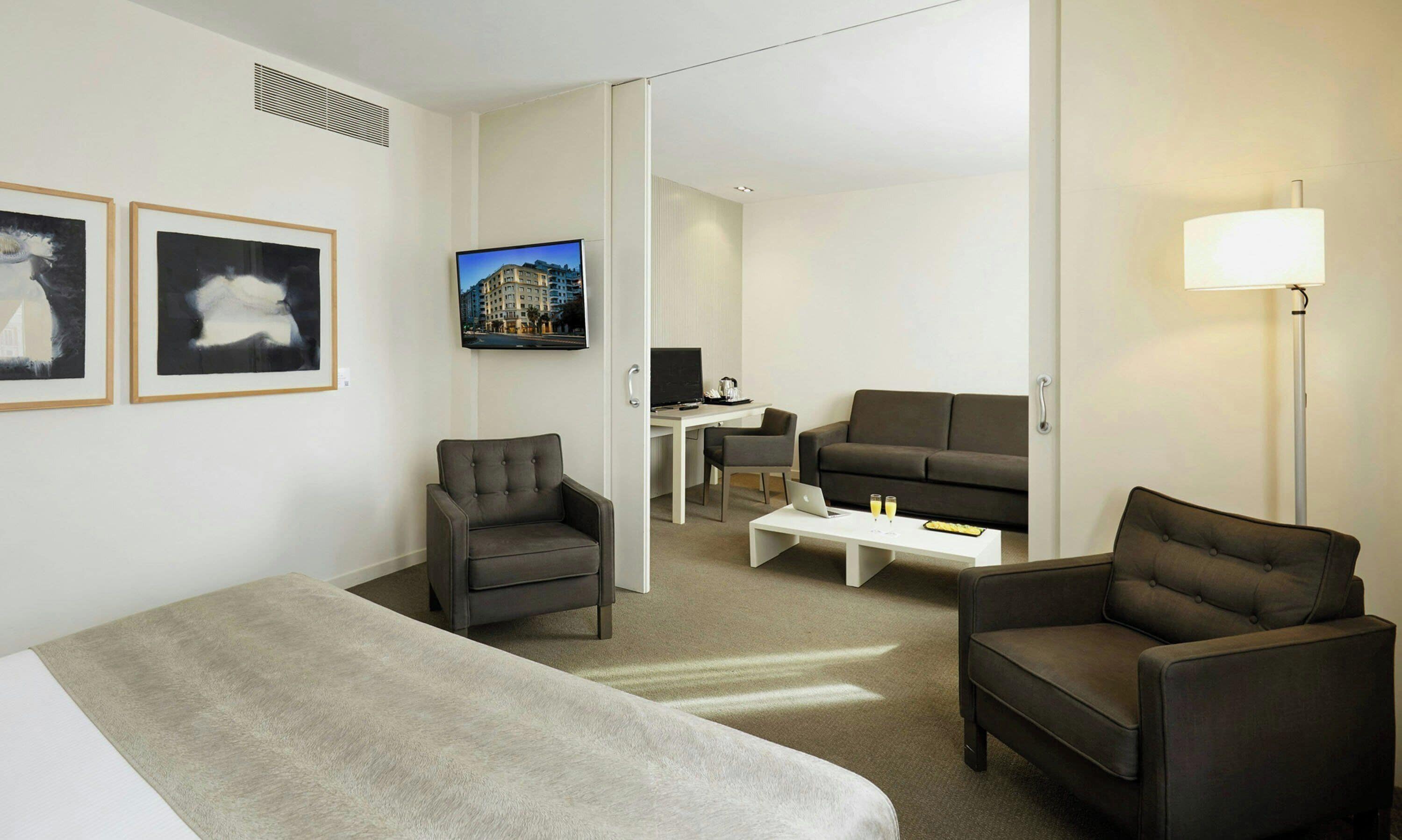 A hotel room with a bed, two armchairs, a sofa, a desk with a computer, a flat-screen TV, and a lamp.