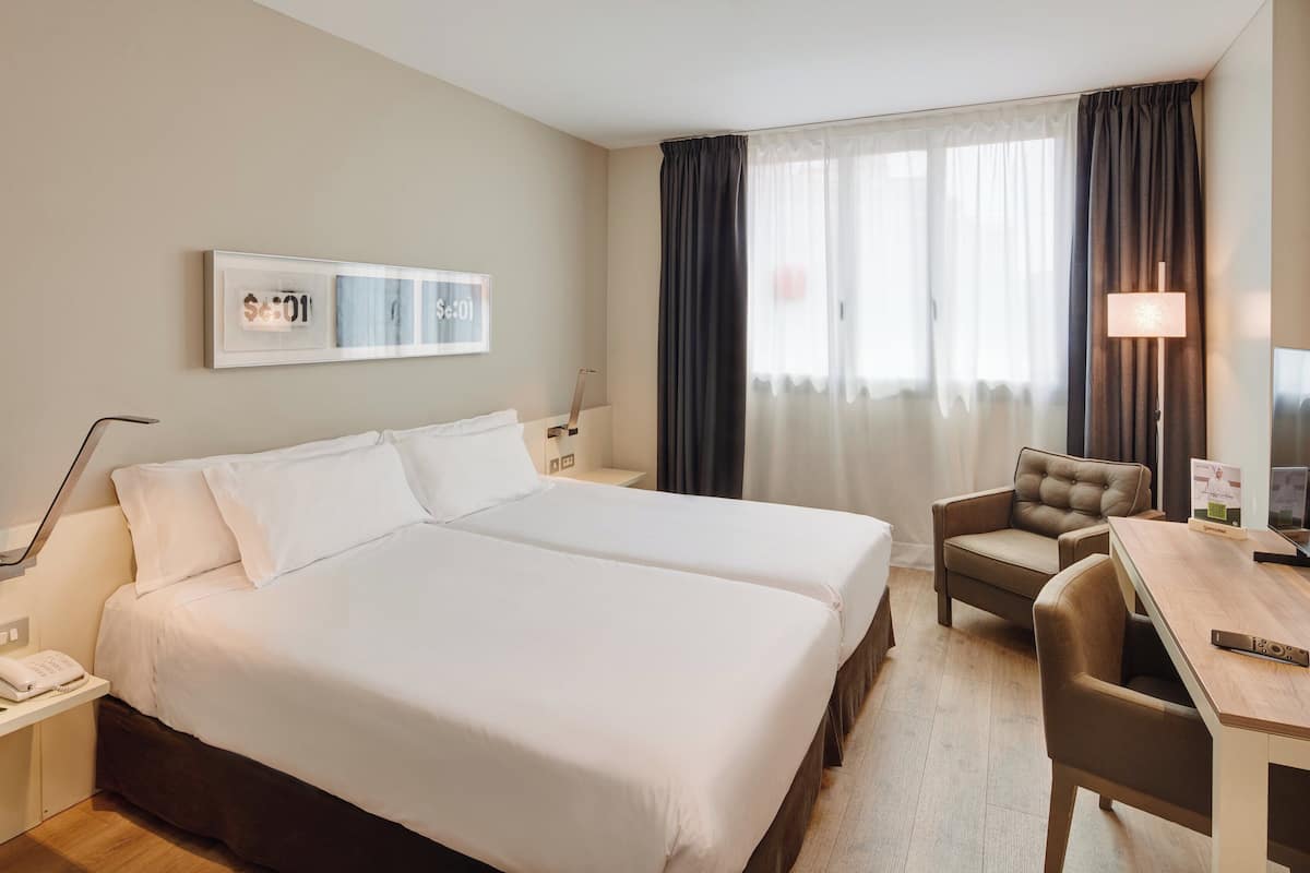 Habitació d'hotel amb dos llits, una taula de treball, una cadira, un televisor i una làmpada.