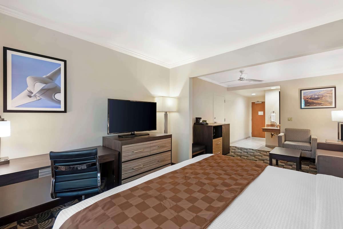 A hotel room with a large bed, a desk with a flat-screen TV, a chair, a dresser, and a small table.