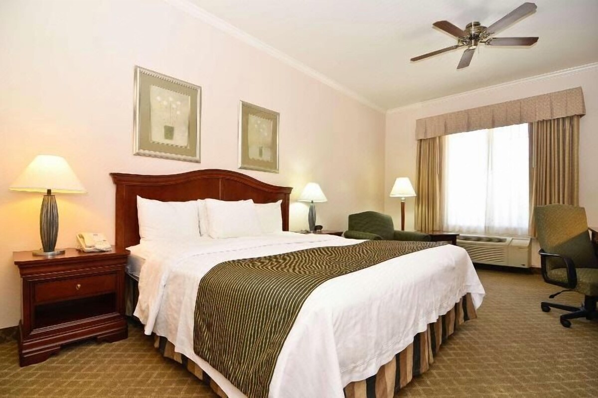 A hotel room with a large bed, two bedside tables, a chair, a desk, a ceiling fan, and a window with curtains.