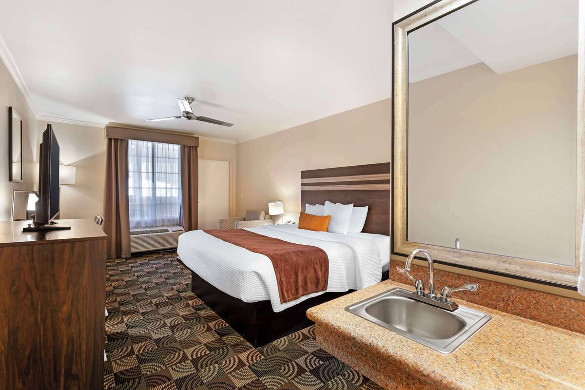A hotel room with a bed, a desk, a large mirror, a sink, and a ceiling fan.