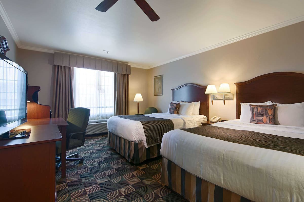 A hotel room with two beds, a desk with a computer, a chair, a window with curtains, and a ceiling fan.
