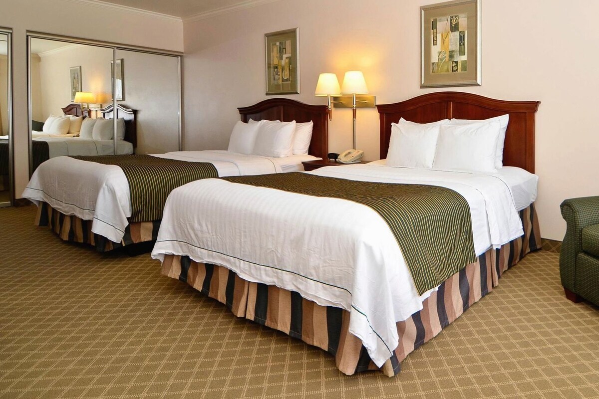 A hotel room with two beds, a mirror, a bedside table with a lamp, and a green chair.