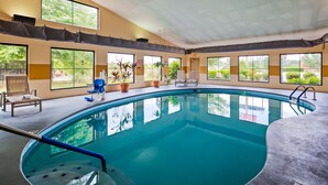 An indoor swimming pool with clear blue water, surrounded by windows offering views of greenery. There are lounge chairs and potted plants around the pool area.