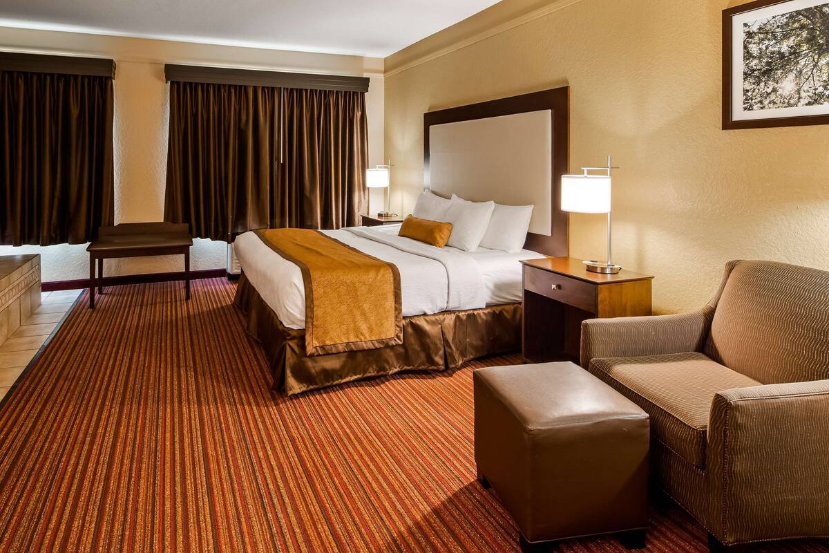 A hotel room with a large bed, a sitting area with a chair and ottoman, a small table, and a bathtub.