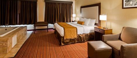 A hotel room with a large bed, a sitting area with a chair and ottoman, a small table, and a bathtub.