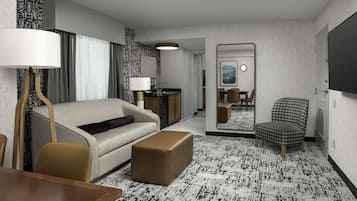 Habitación de hotel con sofá, reposapiés, mesa de comedor y sillas. Cuenta con un espejo, una lámpara y una pequeña kitchenette.
