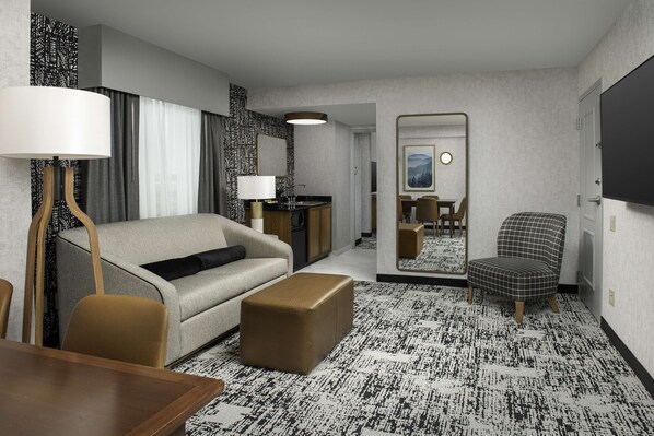 Habitación de hotel con sofá, reposapiés, mesa de comedor y sillas. Cuenta con un espejo, una lámpara y una pequeña kitchenette.