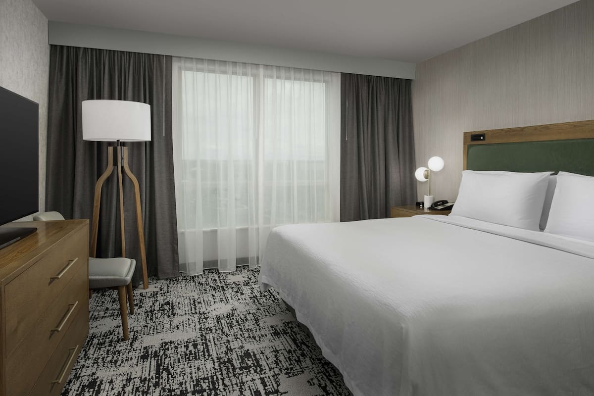 Habitación de hotel con una cama grande, un escritorio con lámpara, una silla y vistas al exterior a través de cortinas translúcidas.