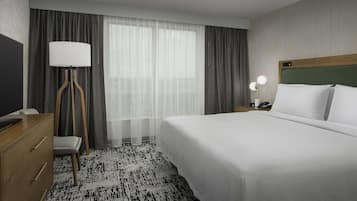 A hotel room with a large bed, a desk with a lamp, a chair, and a view of the outdoors through sheer curtains.