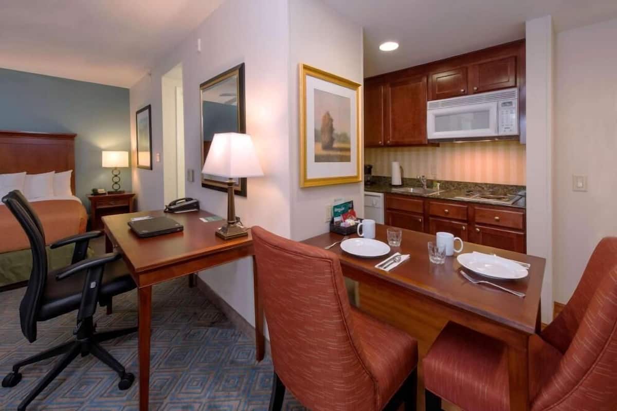 A hotel room with a bed, desk, chairs, a lamp, a microwave, and a kitchen area.