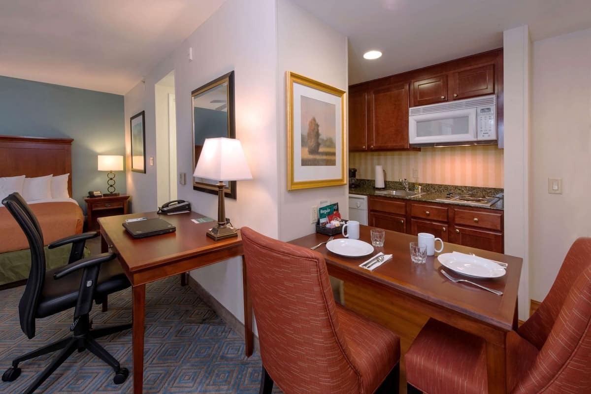 A hotel room with a bed, desk, chairs, a lamp, a microwave, and a kitchen area.