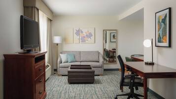 A hotel room with a television, sofa, ottoman, desk, chair, and artwork on the walls.