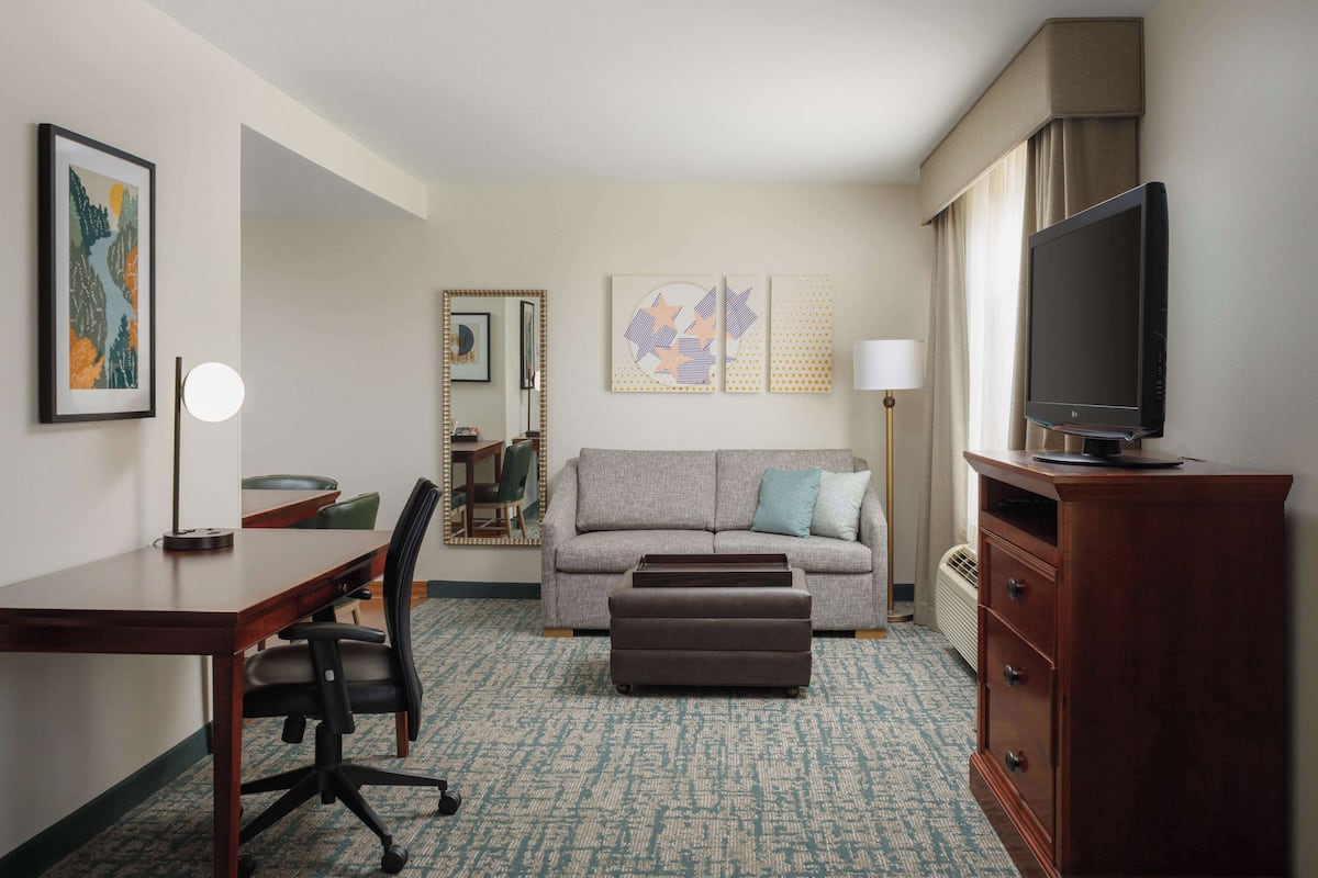 A hotel room with a desk, office chair, sofa, ottoman, TV, and a lamp.
