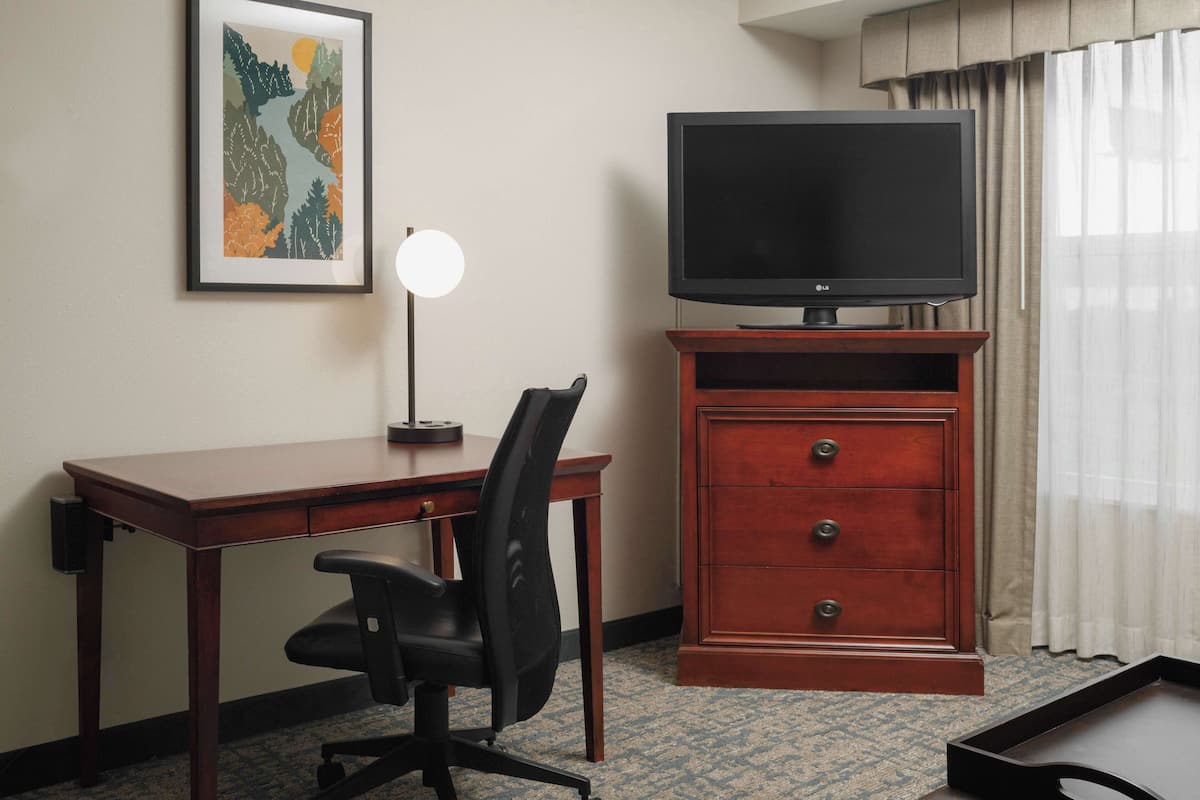 A hotel room with a wooden desk, a black office chair, a television on a wooden stand, a framed picture on the wall, and a window with curtains.