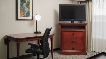 A hotel room with a wooden desk, a black office chair, a television on a wooden stand, a framed picture on the wall, and a window with curtains.