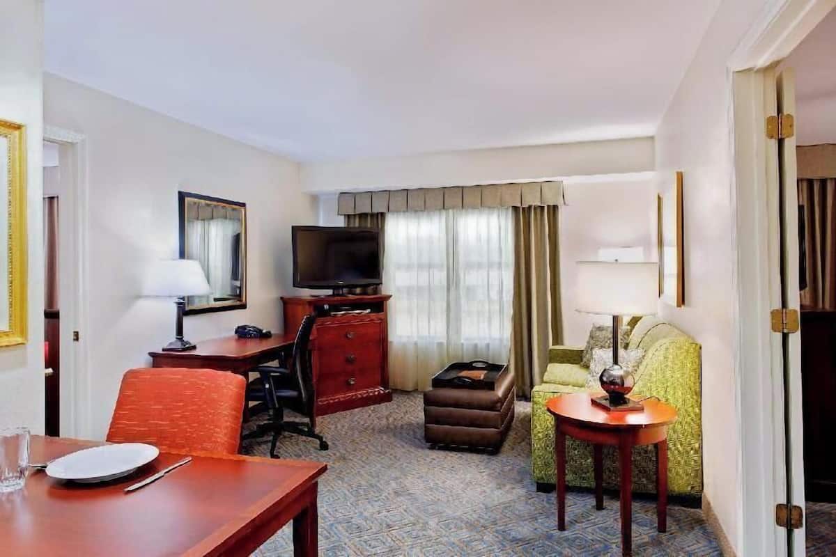 A hotel room with a dining table, a television, a sofa, and a small table with a lamp.